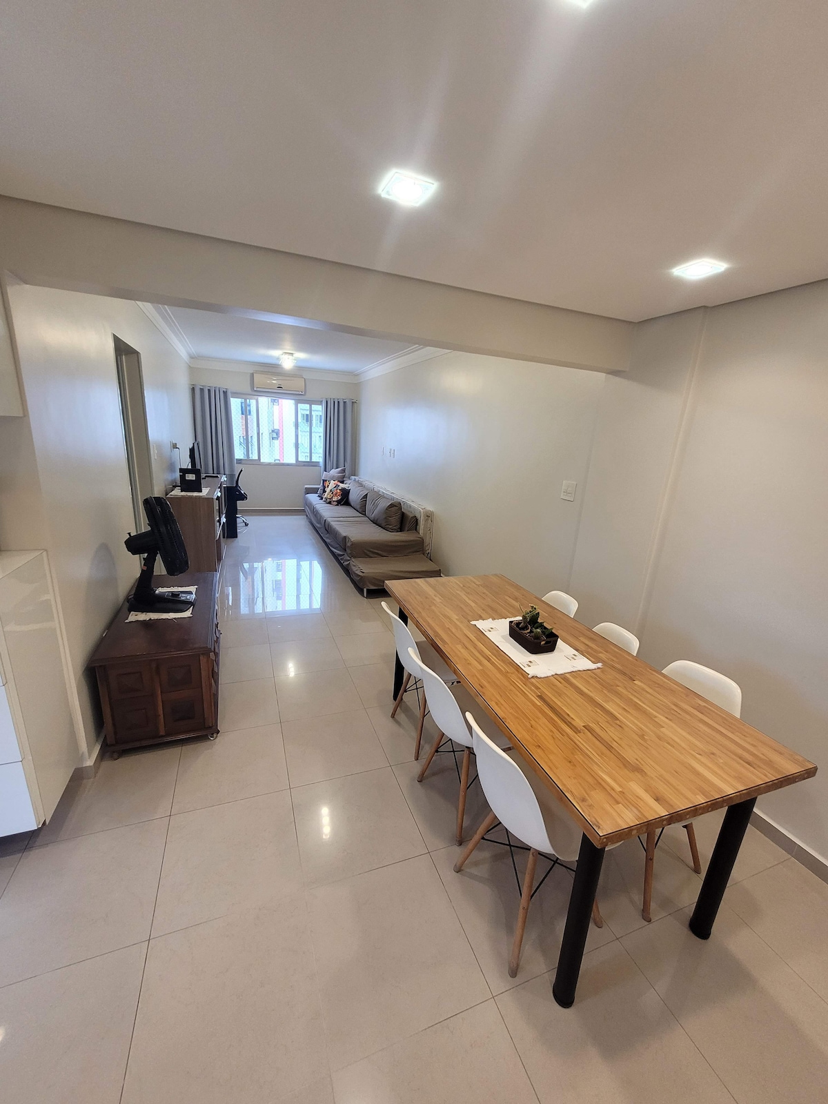 A spacious living area is highlighted by a wooden dining table surrounded by six chairs. A comfortable sofa is positioned nearby, facing a window that allows natural light to illuminate the space. Neutral-colored walls and tiled flooring contribute to the open feel.