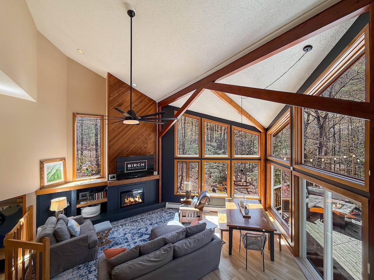 The spacious living area features cathedral ceilings and large floor-to-ceiling windows that invite natural light. A deep sectional sofa is centered in front of an electric fireplace, complemented by a large TV and a dining table positioned beside a window overlooking the serene forest.