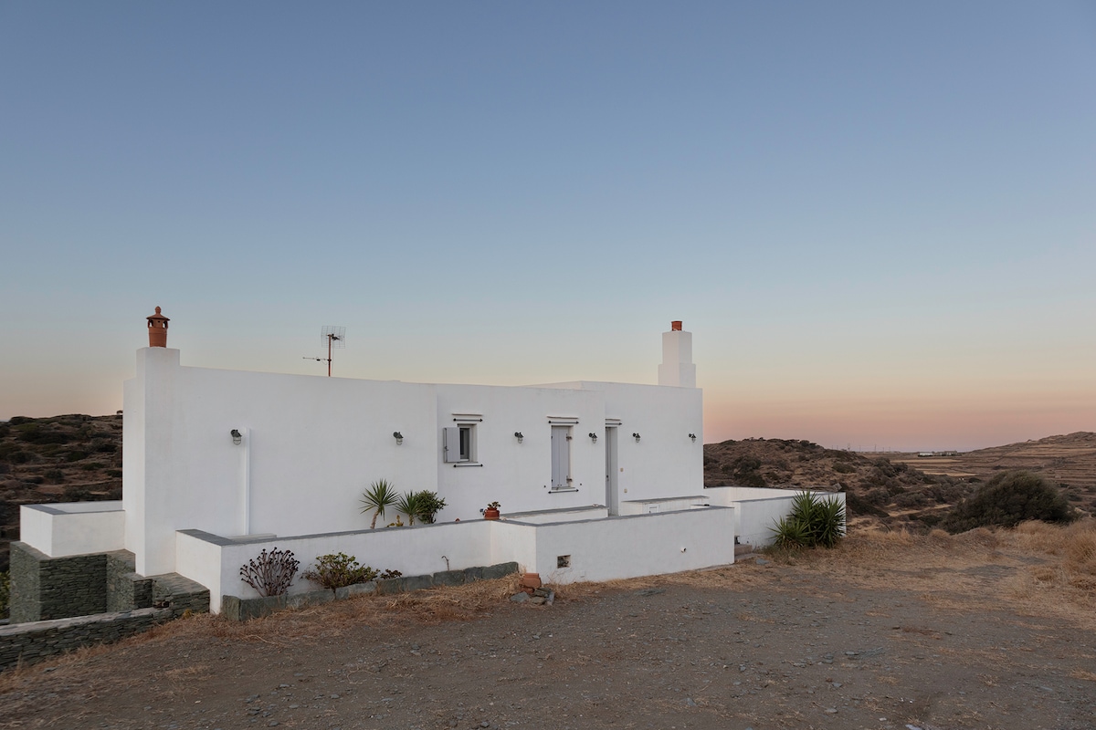A white-walled house is seen against a pastel sunset sky, blending harmoniously with the surrounding natural landscape. The structure features windows and a chimney, with desert-like vegetation surrounding the property. Soft hills can be seen in the background, providing a serene backdrop.