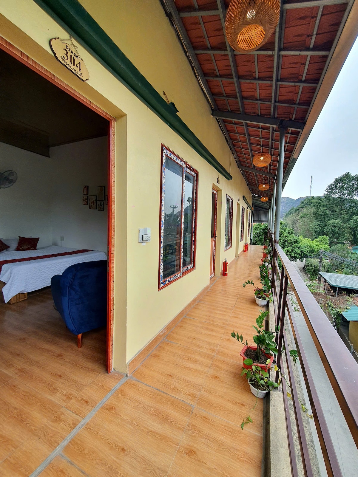 An outdoor corridor is lined with potted plants and features several doors leading into individual rooms. The flooring is tiled in a warm wood tone, while the ceiling showcases decorative light fixtures. The view includes lush greenery visible in the background.