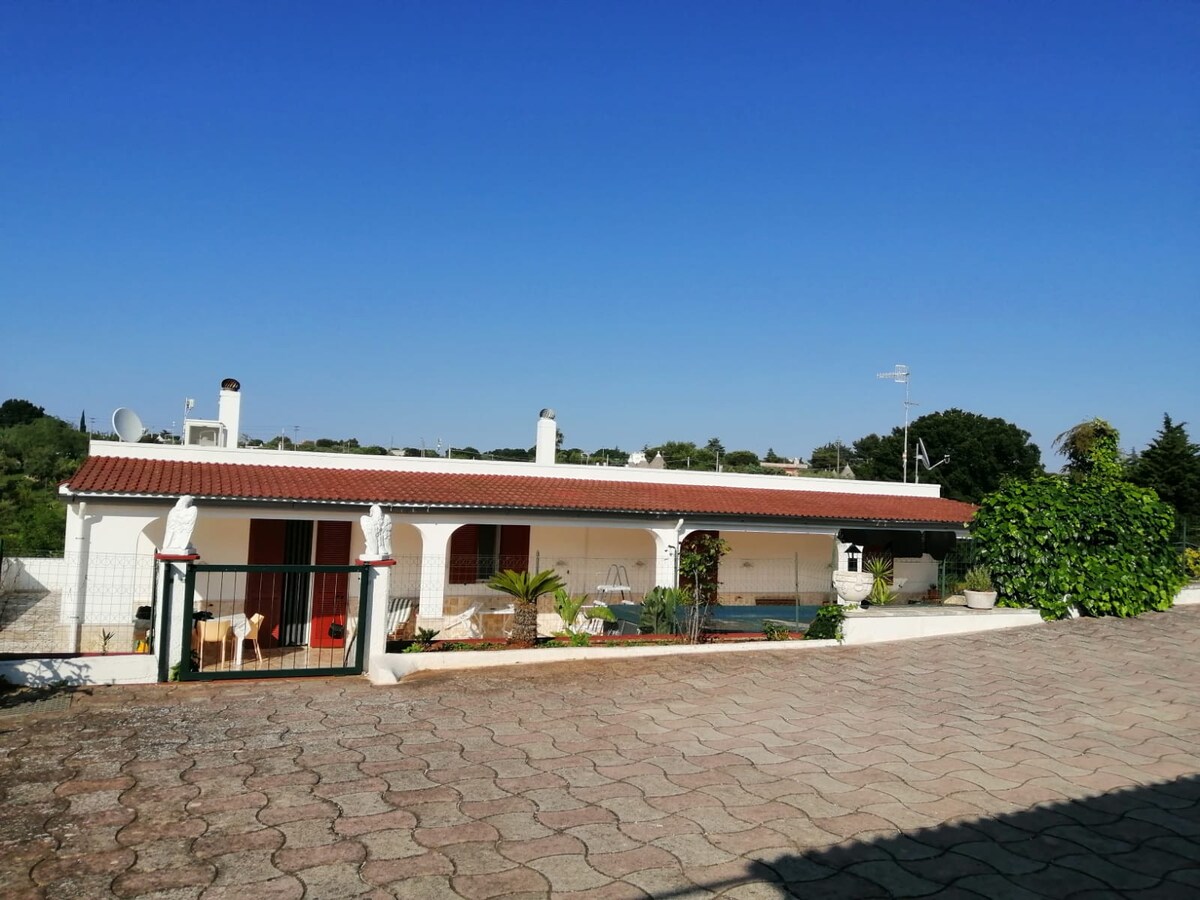 A single-story villa is shown with a terracotta-tiled roof and a wide terrace in front. The structure is surrounded by greenery, and glimpses of patio furniture can be seen. A clear blue sky complements the inviting outdoor space.