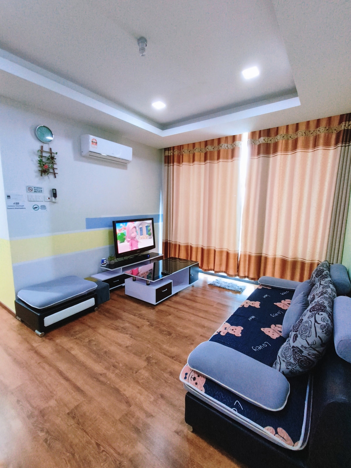 A cozy living room features a comfortable sofa with patterned cushions and a modern television mounted on the wall. Light from large windows filters through soft curtain panels, illuminating the wooden floor. Air conditioning is provided for a pleasant indoor climate.