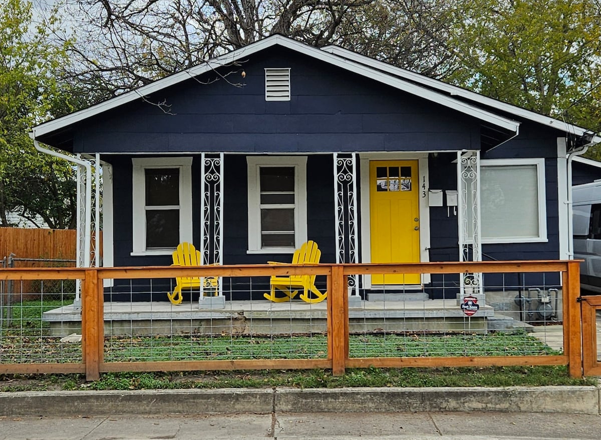 A charming guest house exterior features a navy blue facade with white accents and decorative details. Two bright yellow chairs are positioned on the covered porch, inviting relaxation. A wooden fence encloses a small grassy area in front, enhancing the property's welcoming appearance.