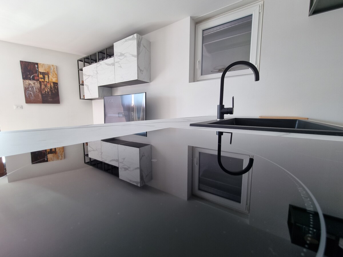 The modern kitchen features a sleek island countertop that reflects the surroundings. Light is introduced through a nearby window. Minimalist cabinetry in light tones is visible, alongside stainless steel appliances, including a refrigerator. A black faucet adds a contemporary touch at the sink area.