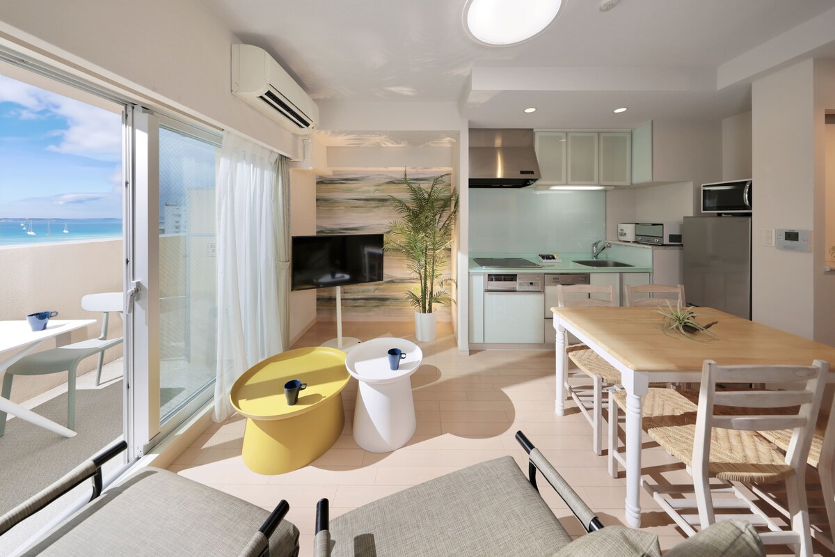 An open living space features a modern kitchen area and a dining table with chairs. Bright, airy walls complement two colorful side tables and a television, while large windows provide a view of the outdoors. Natural light floods in, enhancing the welcoming environment.