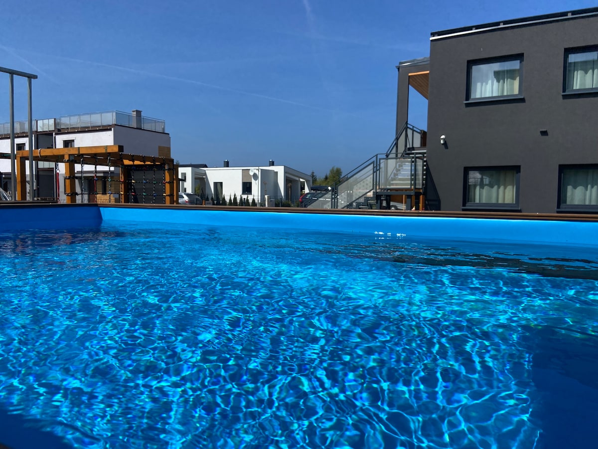 A modern outdoor pool features clear, sparkling water under a bright blue sky. Nearby buildings are visible, showcasing a contemporary architectural design. The pool's edge is surrounded by a wooden deck, providing an inviting space for relaxation.
