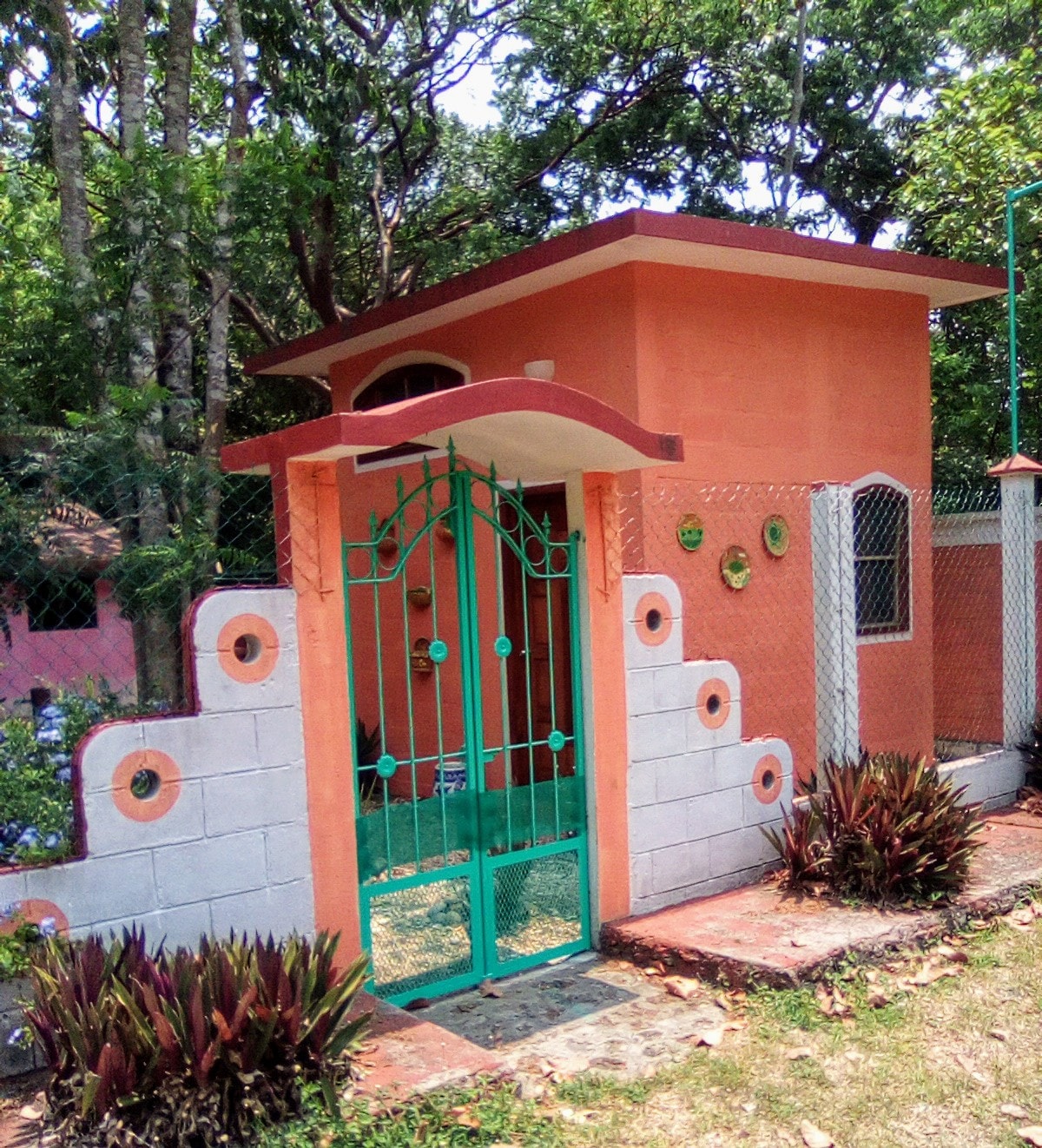 A vibrant orange entrance gate is framed by a decorative wall featuring circular designs in various colors. Lush greenery and colorful plants are positioned along the pathway, creating an inviting entryway to the property surrounded by abundant foliage.