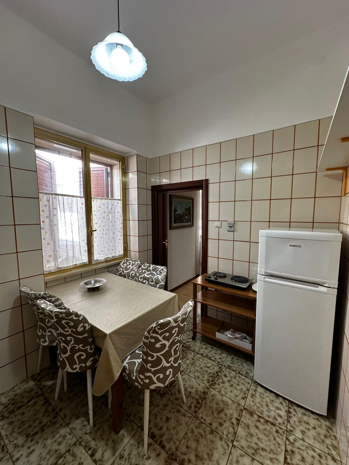 A compact kitchen area features a small dining table with four patterned chairs, accompanied by a fridge and a countertop with kitchen essentials. Tiled walls and a window provide natural light, while a simple light fixture illuminates the space.