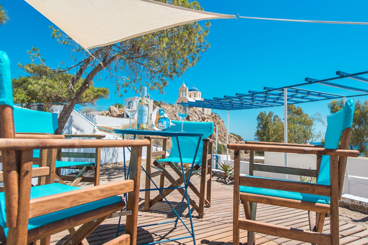 An outdoor seating area features wooden chairs with blue cushions, complemented by a small table. A shaded area provides comfort under the sun, with a view of a distant church atop a rocky hill, surrounded by greenery and a bright blue sky.