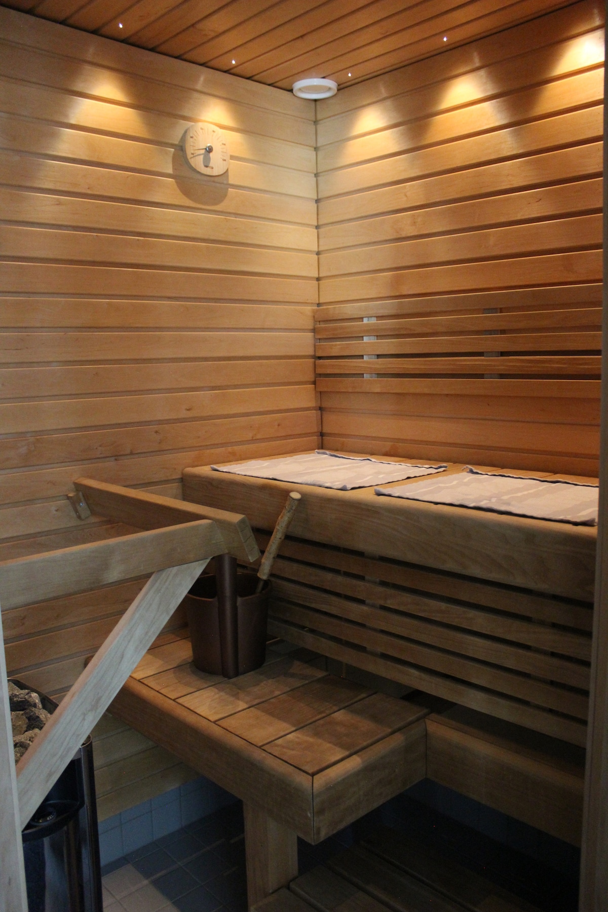 A wooden sauna interior is shown, featuring bench seating along the walls and a slatted wood design. Accessories including a sauna bucket and ladle are positioned on a wooden shelf, while a temperature gauge is mounted on the wall. Soft lighting enhances the natural wood tones.
