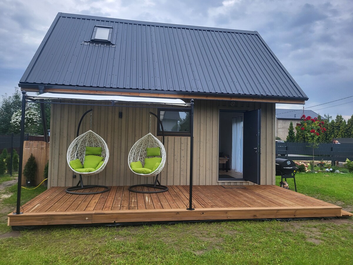 A modern cabin features a welcoming wooden deck, with two green hanging chairs providing a comfortable seating area. The exterior showcases a blend of wood and metal, while a grill is positioned nearby, complementing the grassy surroundings.