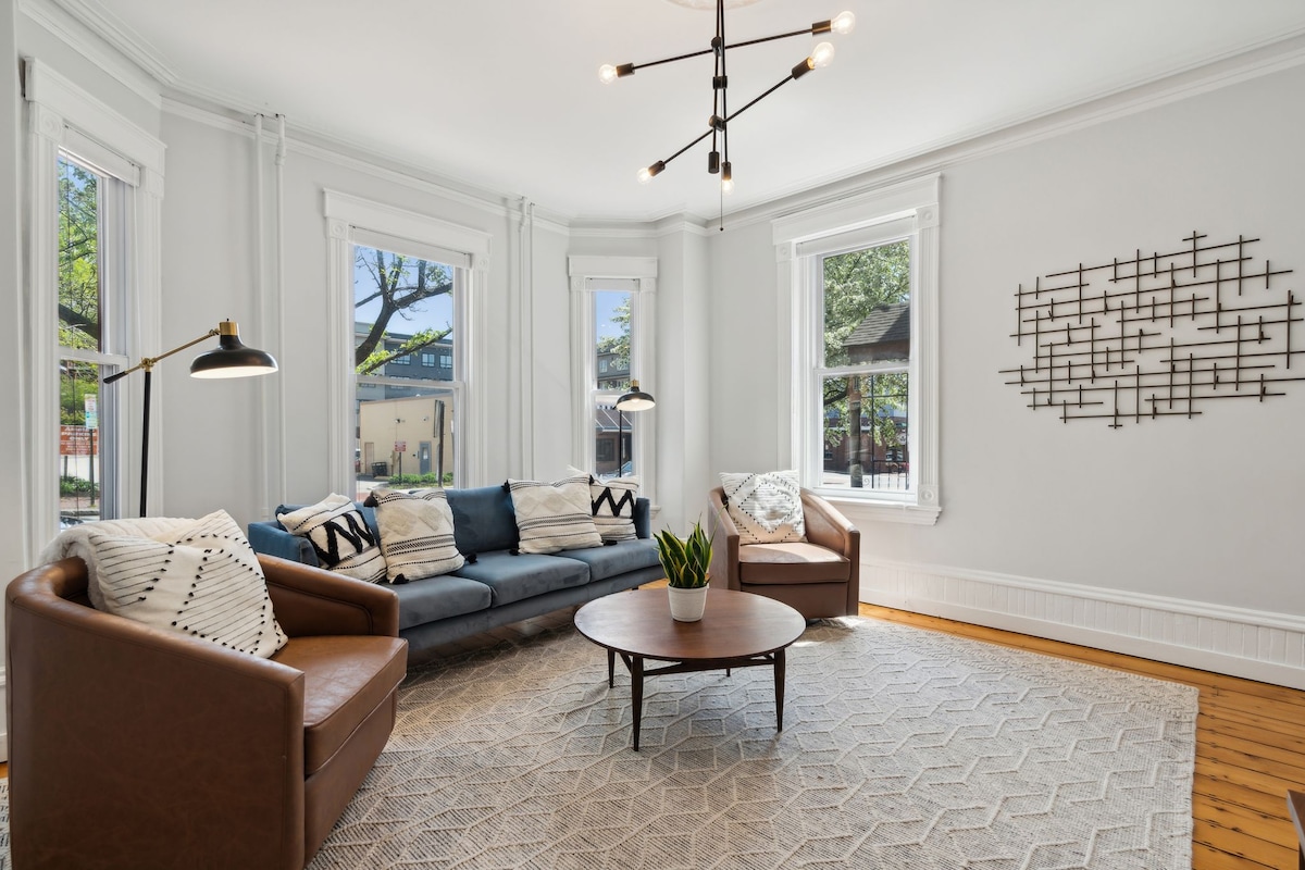 A spacious living room is highlighted by large windows allowing natural light to fill the area. Comfortable seating is provided by a dark sofa and two armchairs. A round coffee table rests on a patterned area rug, with a decorative wall piece adding visual interest.