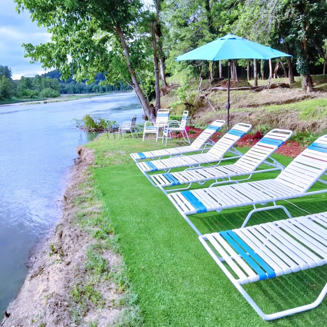 Lawn chairs with striped cushions are arranged by the river, under the shade of a teal umbrella. The grassy area provides a relaxing spot to enjoy the views of the water lined by trees in the background.