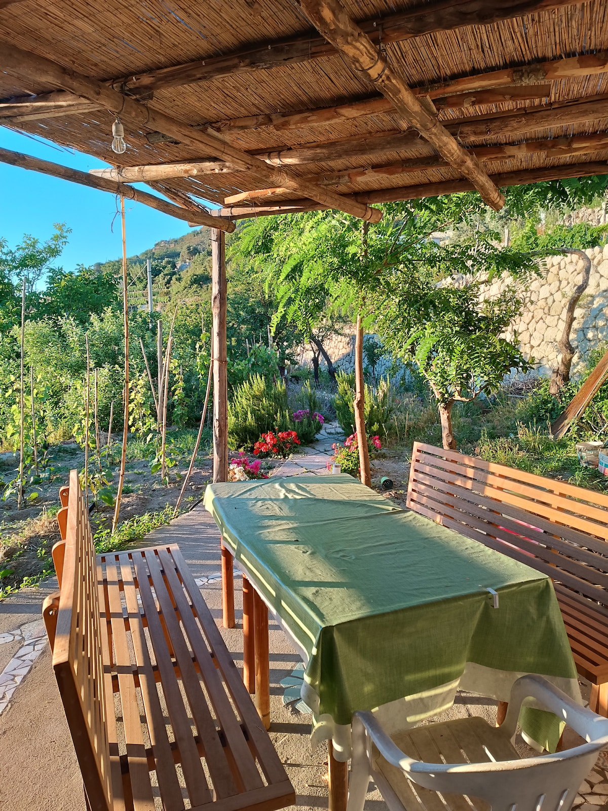An outdoor dining area features a wooden table covered with a green cloth, surrounded by benches. The space is shaded by a thatched roof and overlooks a garden with blooming flowers and greenery, creating a serene environment for meals and relaxation.