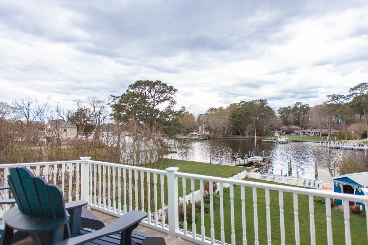 A serene view from the deck showcases the tranquil river, bordered by lush greenery. The gently flowing water reflects the cloudy sky, while several docks are visible along the shoreline. Comfortable chairs are positioned to enjoy the scenery.