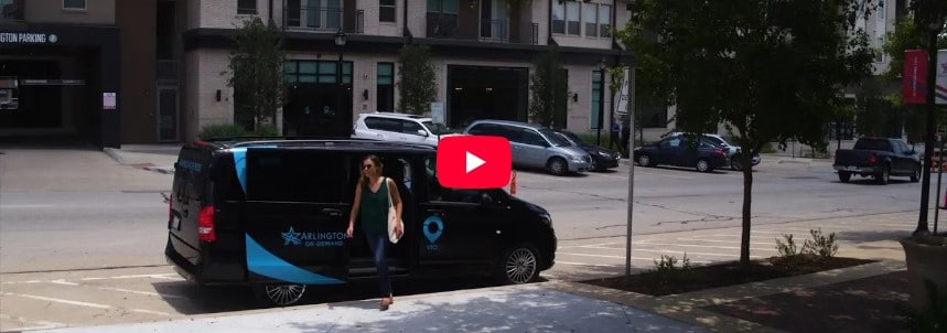 A passenger exits a rideshare van parked along a bustling street. Surrounding the vehicle, a variety of cars is visible, set against the backdrop of modern apartment buildings. Green trees frame the scene, contributing to a lively urban environment.