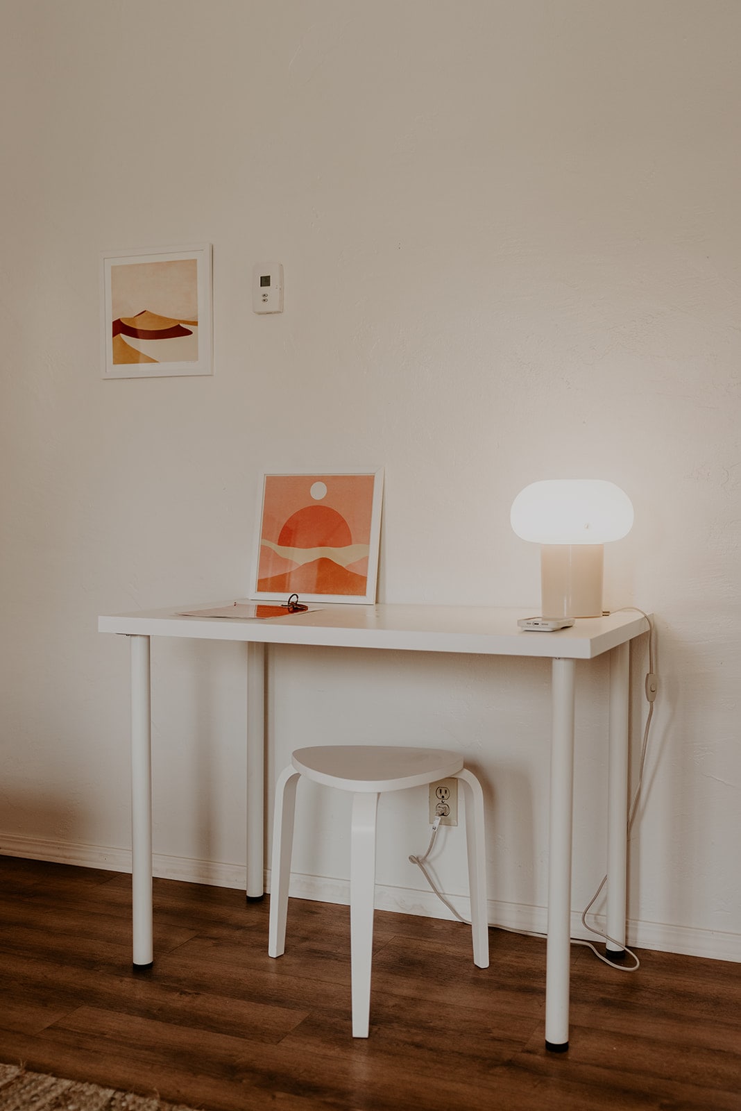 A clean and minimal workspace is depicted, featuring a simple white table and stool. A soft-glowing lamp and framed artwork add a touch of color to the wall. The warm wood flooring complements the light tones of the furniture.