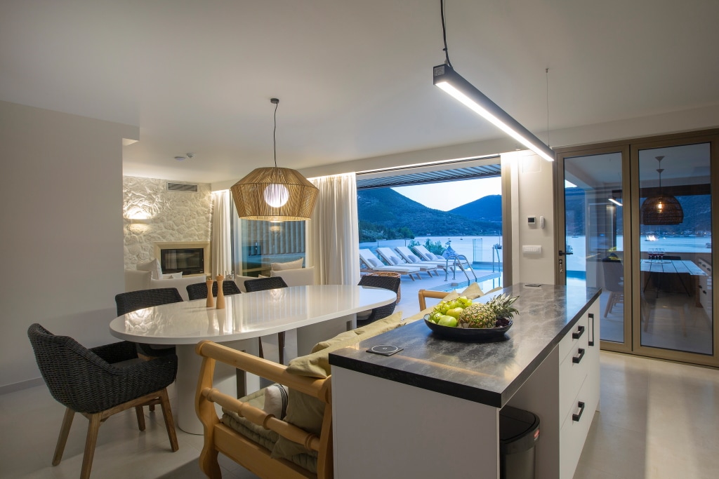 An inviting dining area features a large round table surrounded by woven chairs. Natural light pours in through expansive sliding glass doors, revealing stunning views of the outdoor pool terrace and mountains beyond. A contemporary pendant light hangs above the table, enhancing the spacious feel.