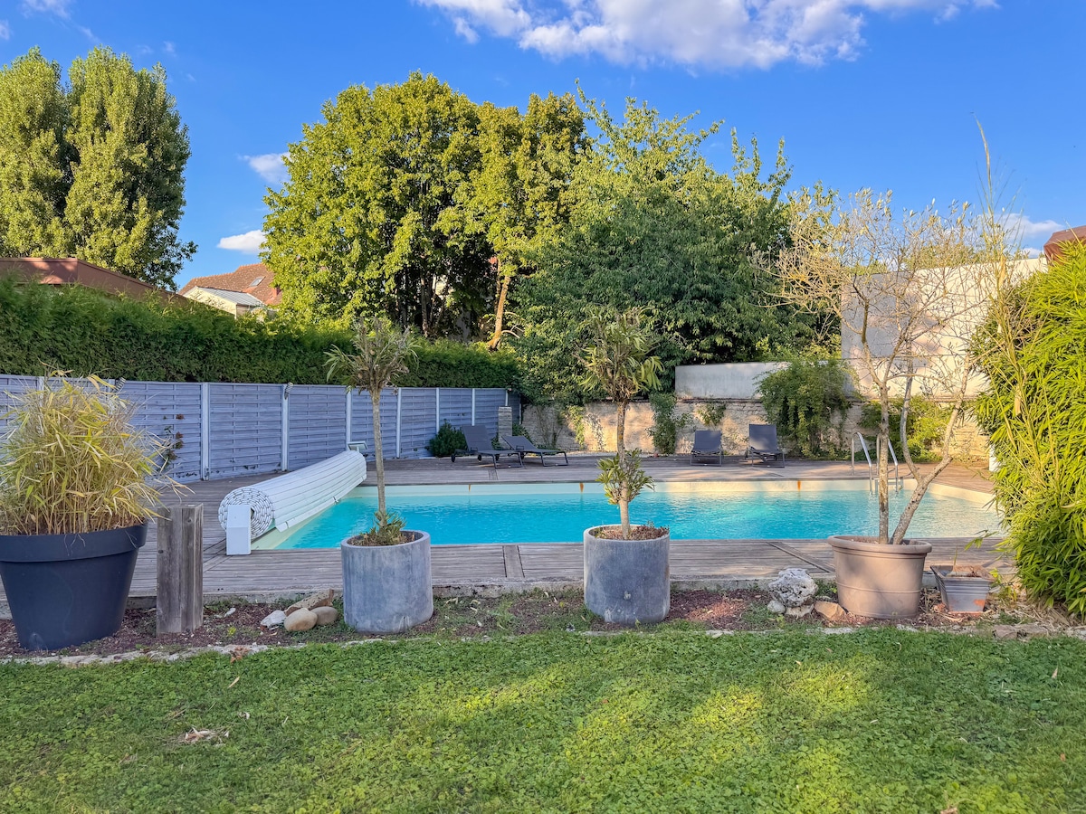 A tranquil outdoor space features a large swimming pool surrounded by greenery and flowering plants. Potted plants are arranged along the poolside, and sun loungers are positioned for relaxation. The clear blue sky is complemented by trees providing shade to the area.