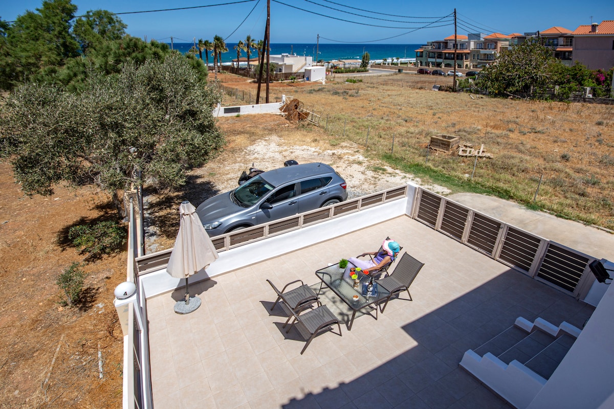 An outdoor terrace features a table set for outdoor dining, surrounded by two lounge chairs. The well-maintained area is complemented by a patio umbrella. A view of the sea is visible in the background, along with a grassy field and parked car.