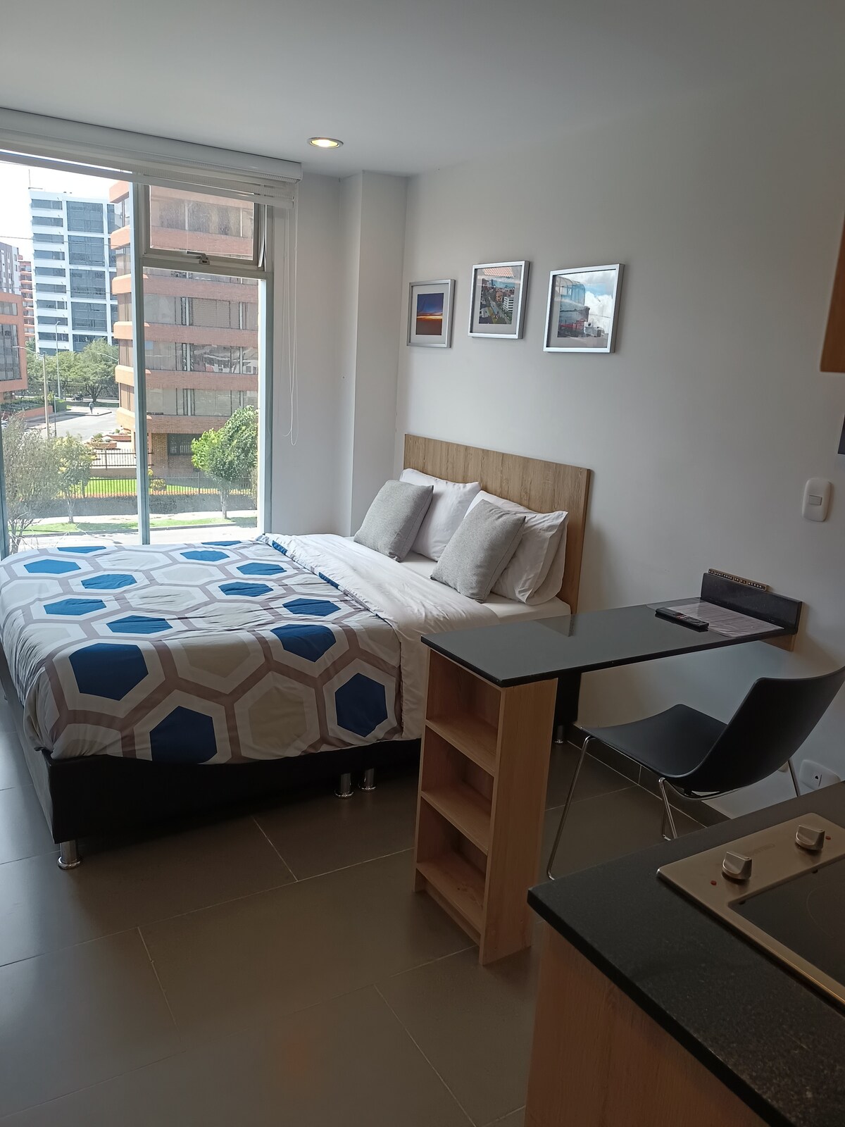 A cozy sleeping area features a spacious bed with a patterned blanket, complemented by light-colored pillows. A small kitchen bar with stools provides a dining space. A window allows natural light to illuminate the room, with views of the exterior greenery and buildings.