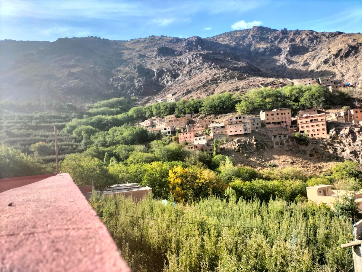 A scenic view of a mountainous landscape featuring a series of buildings nestled among lush greenery. The hills rise dramatically in the background, while a mix of trees and residences forms a harmonious blend of nature and architecture.