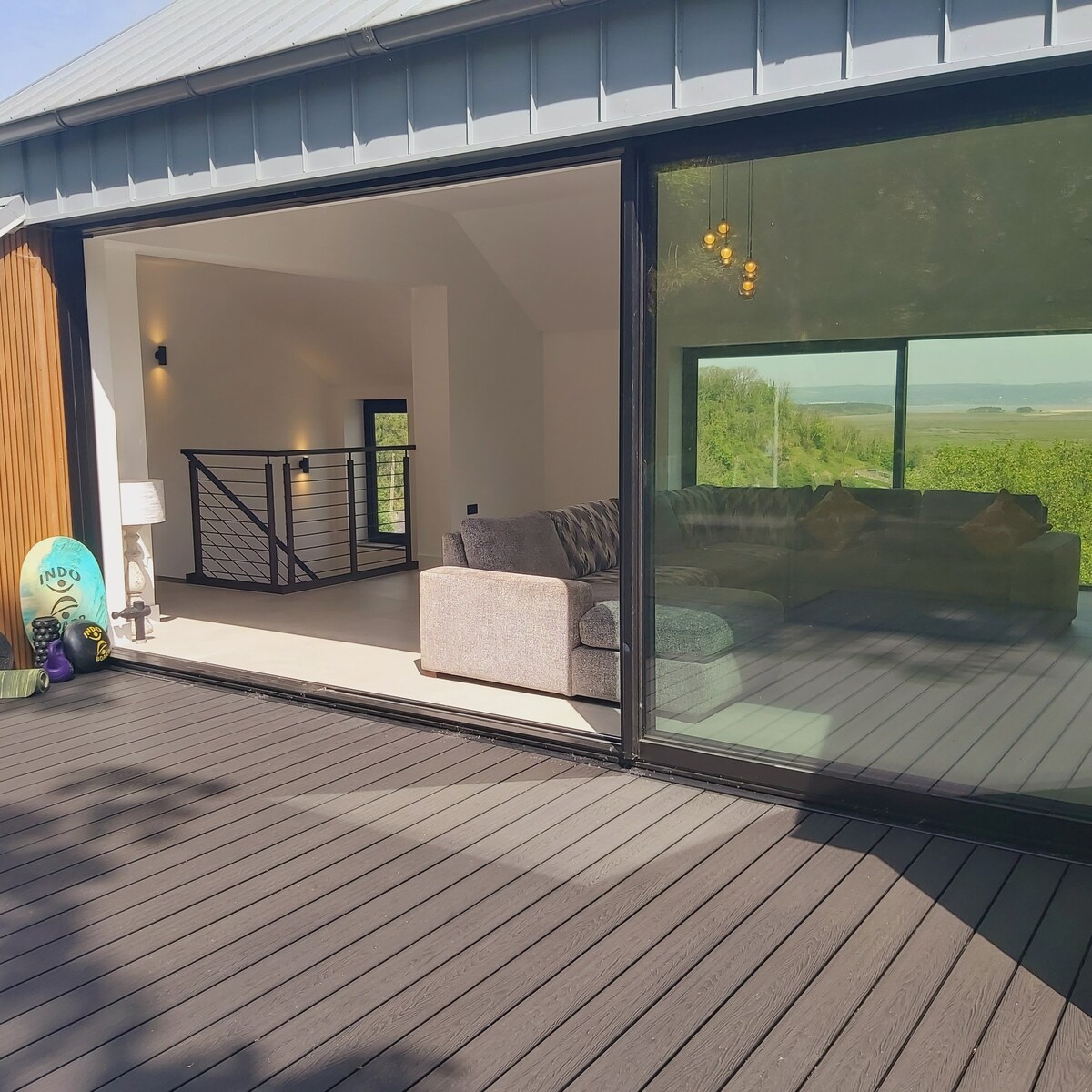A modern outdoor deck features a large sliding glass door leading to an open living space. A spacious sectional sofa faces the door, while views of the surrounding greenery can be seen beyond. The deck showcases a smooth wooden surface and a minimalistic railing.