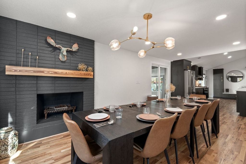 An elegant dining area is presented with a large dark wood table set for eight. Stylish leather chairs surround the table. A mounted decoration resembling antlers is positioned above a minimalist fireplace, while a modern light fixture hangs overhead, adding a touch of sophistication.