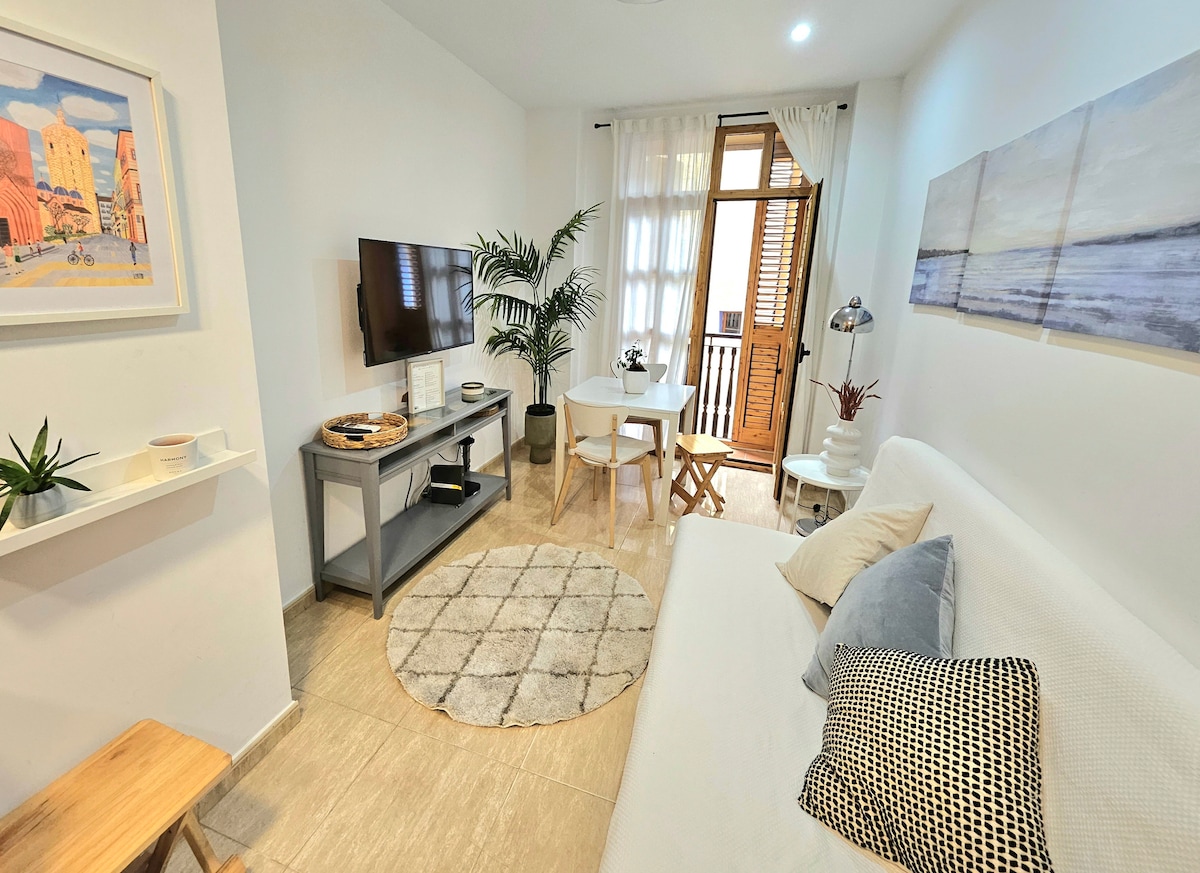 A cozy living space is shown with a neutral color palette. A white sofa is positioned beside a round decorative rug. A dining area features a table and two chairs, while a flat-screen television is mounted on the wall. Natural light enters through a window adorned with wooden shutters.