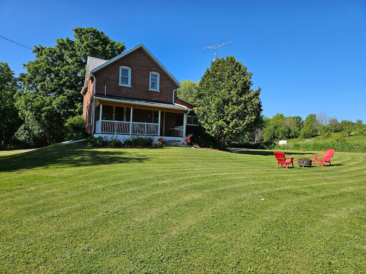 A spacious brick house is set in a lush green landscape. A cozy porch invites relaxation, while two red chairs are positioned by a small fire pit on the lawn. Surrounding trees provide some shade, and the sky above is clear and bright.