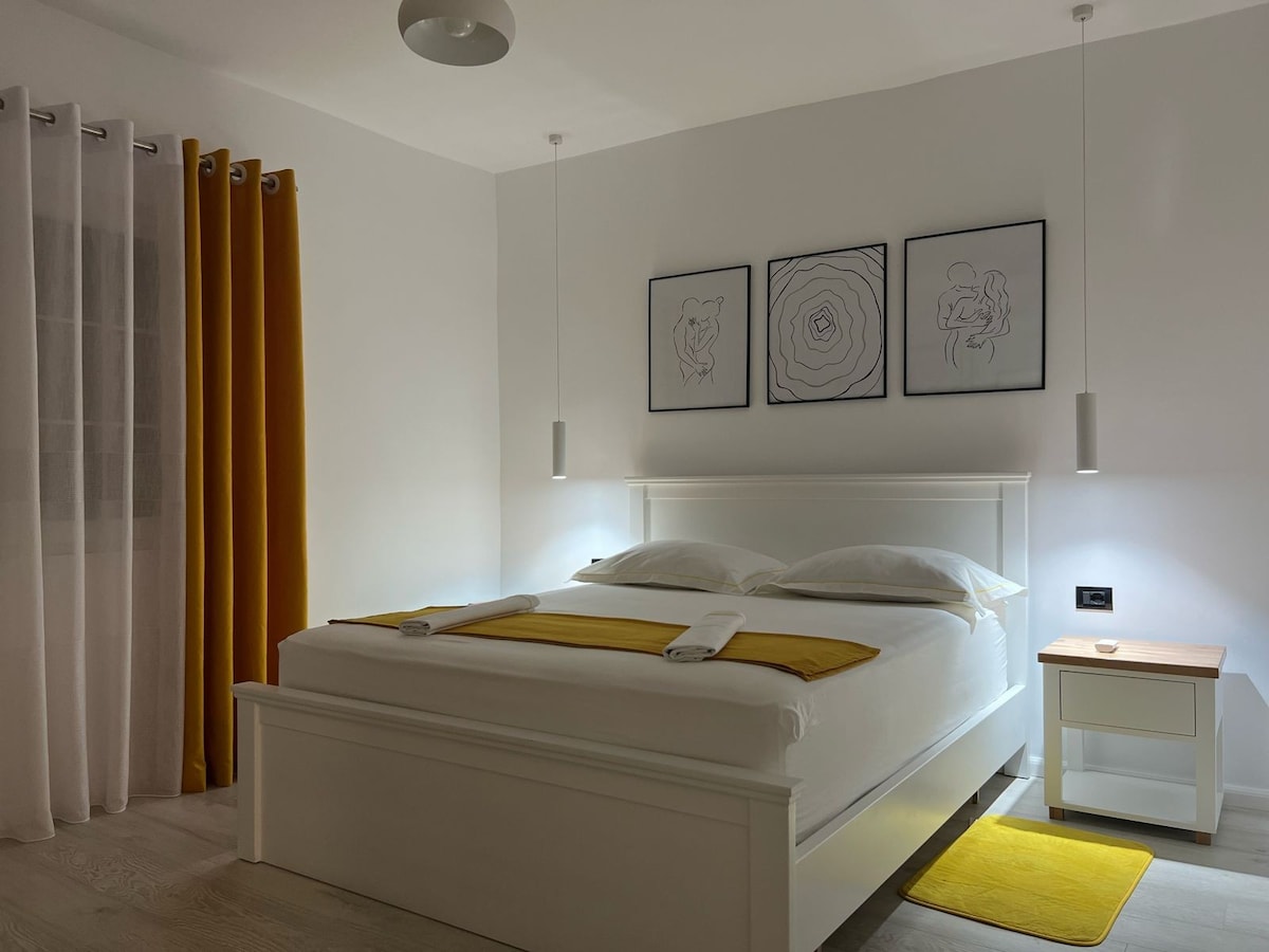 A well-lit bedroom features a spacious bed with white linens and a mustard yellow accent blanket. Two modern bedside lamps provide soft illumination, and artwork is displayed above the bed. Floor-to-ceiling curtains in light gray complement the mustard yellow curtains, creating a balanced color scheme.
