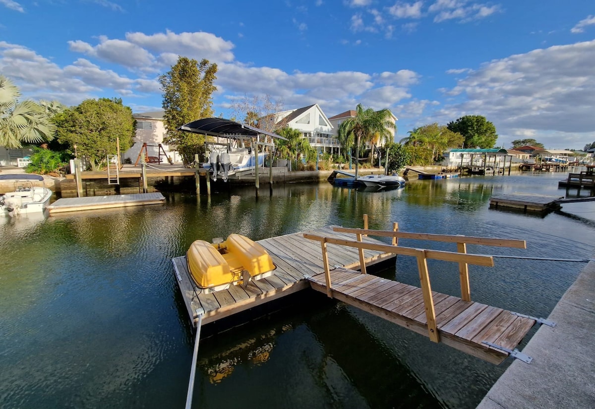 A private dock extends over calm waters, featuring two yellow kayaks resting alongside. Lush greenery and nearby boats create a peaceful waterfront scene. The sky is partly cloudy, allowing natural light to reflect off the water's surface.