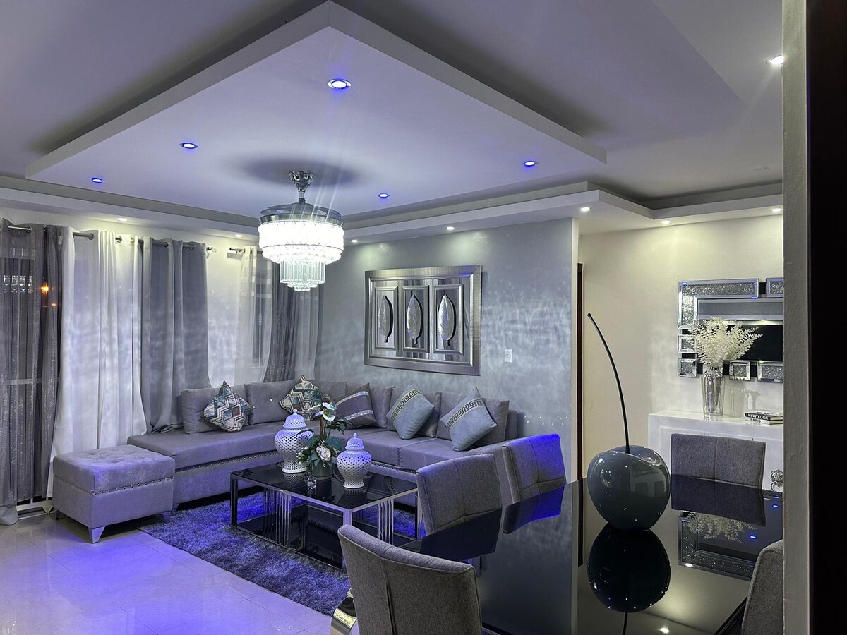 A modern living area is depicted, featuring a large sectional sofa accentuated with decorative cushions. A glass coffee table sits centrally, surrounded by stylish chairs. Ambient lighting is provided by a contemporary chandelier and built-in ceiling lights, highlighting the elegant decor and spaciousness.