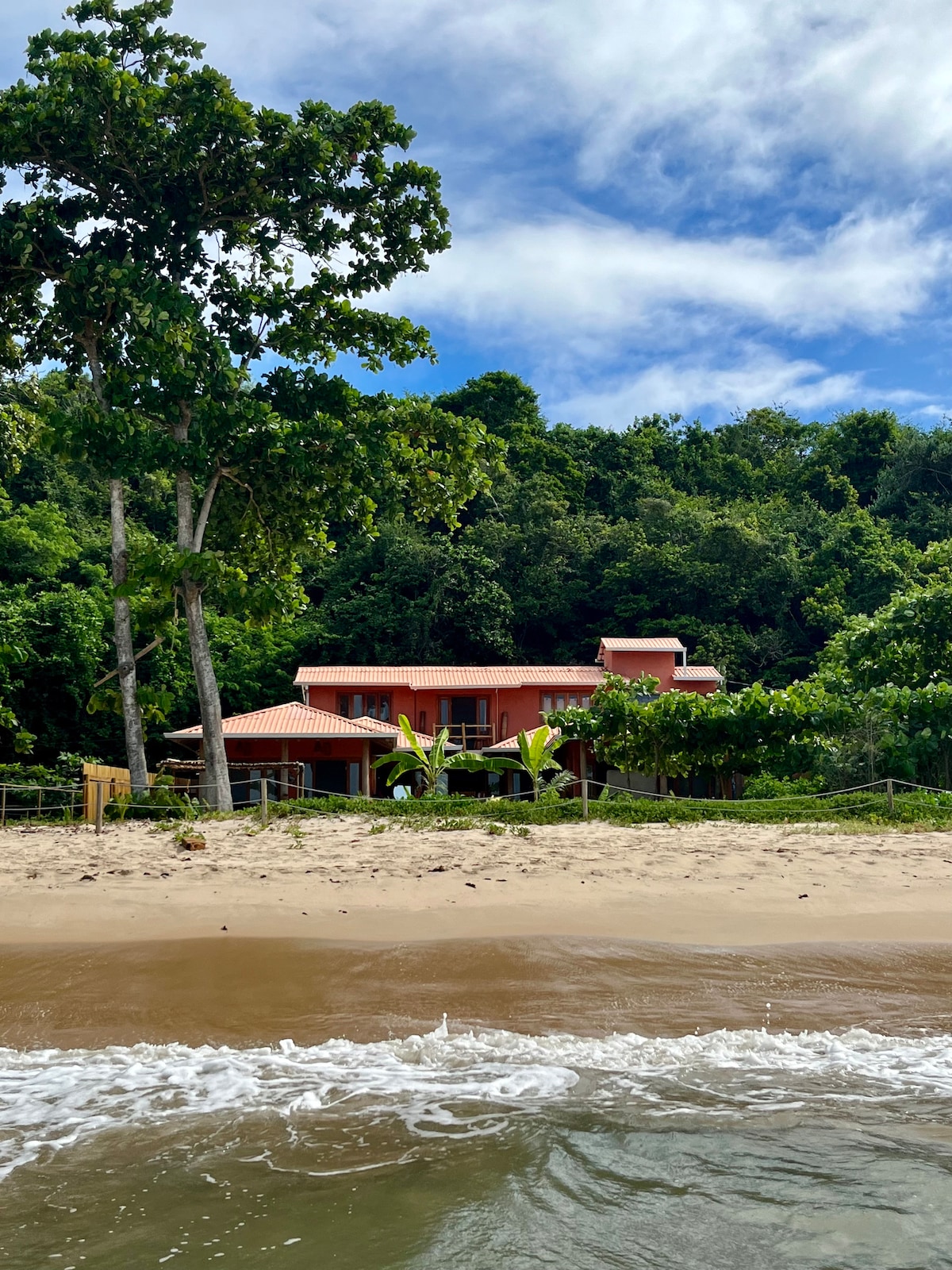 A beachside house is set amidst lush greenery, showcasing its vibrant coral exterior. The expansive structure features multiple levels and a welcoming terrace. Gentle waves lap at the sandy shore in the foreground, enhancing the serene coastal environment.