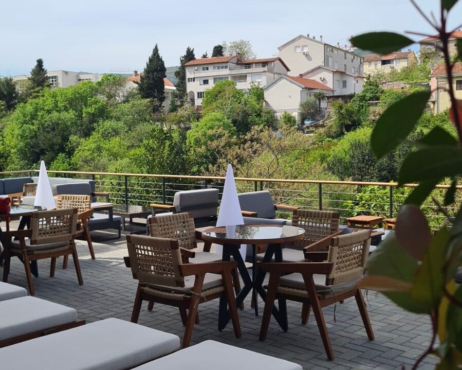 An outdoor seating area features wooden chairs and tables arranged on a paved terrace. A backdrop of lush greenery and residential buildings is visible in the distance. White tablecloths and napkins are elegantly placed on the tables, creating an inviting atmosphere for social gatherings.