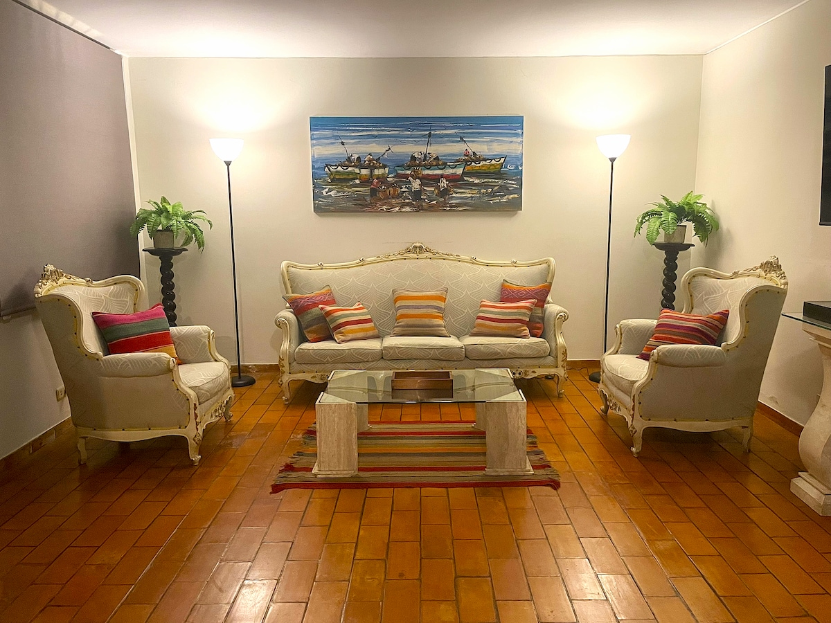 A comfortable living area features a cream-colored sofa with colorful striped cushions and ornate detailing. Flanking the sofa are two elegant armchairs, and a glass coffee table sits on a striped rug. Soft lighting is provided by two standing lamps, with greenery adding a touch of color.