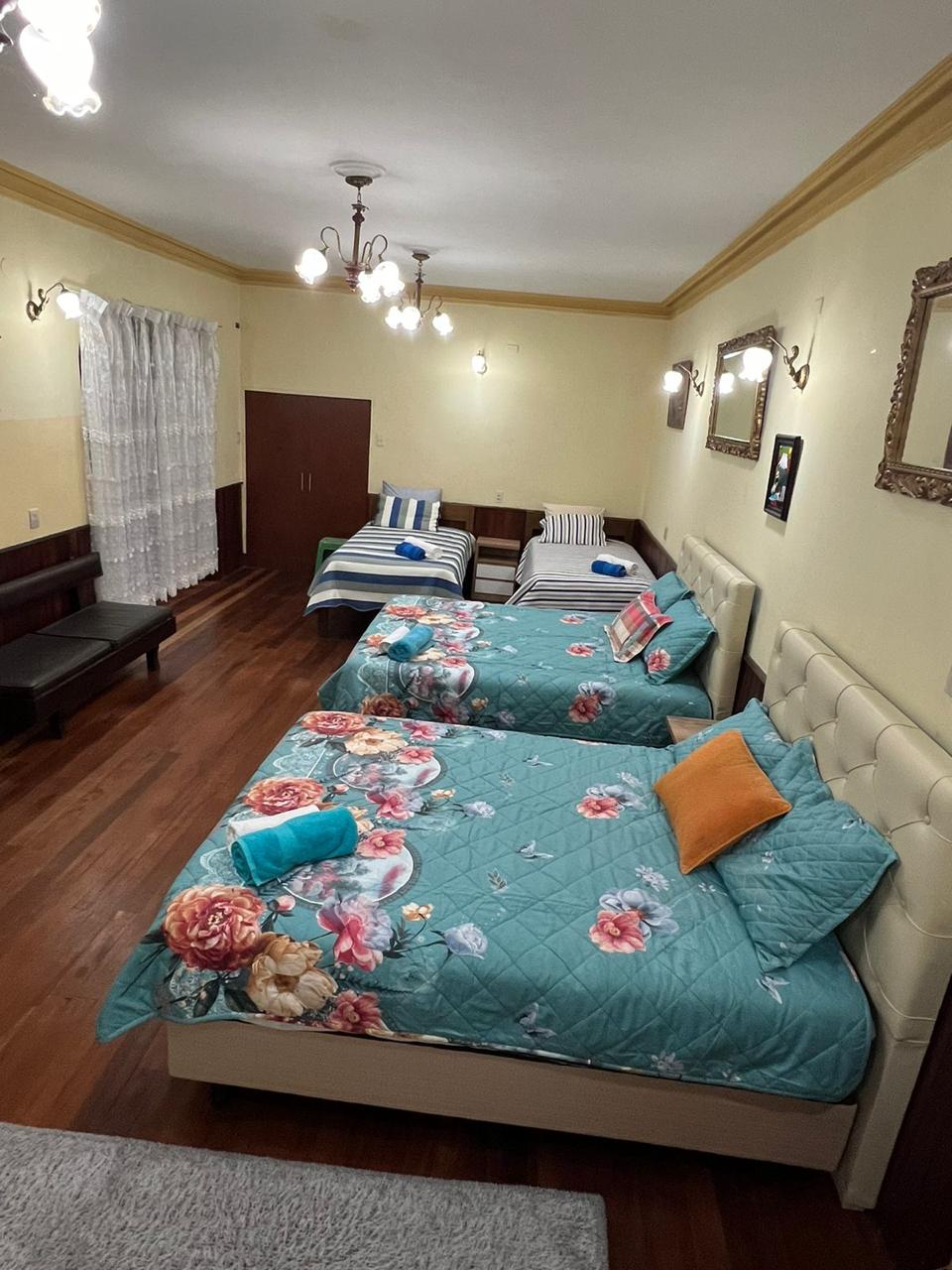 A spacious bedroom is furnished with two larger beds and two smaller beds, all adorned with floral bedding. Light wood flooring complements the soft lighting from the ceiling fixtures. A seating area is visible, featuring dark wood benches. Curtains frame the window, enhancing the natural light.