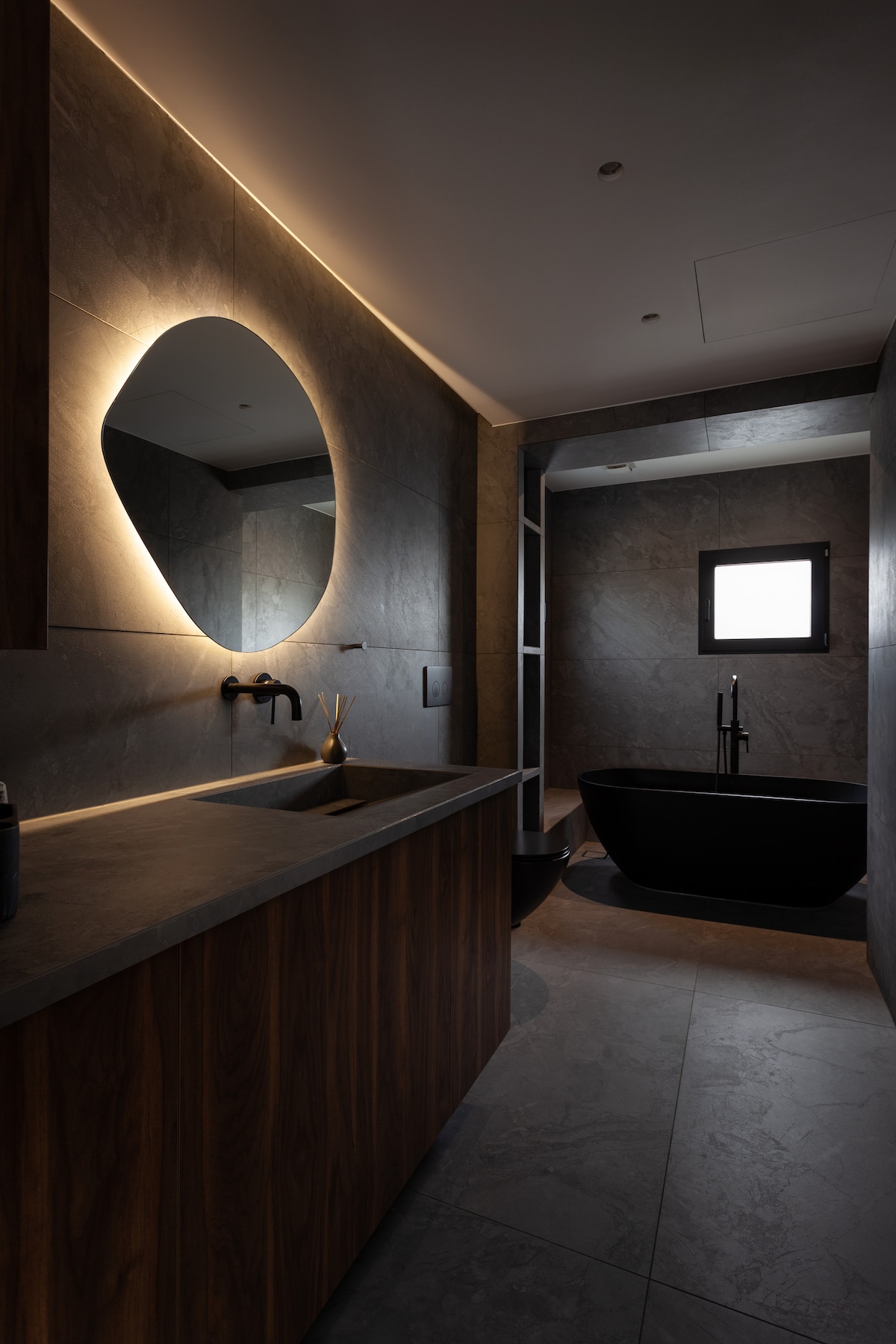 A modern marble bathroom features a sleek wooden vanity with a stylish vessel sink. Ambient lighting highlights an oval mirror above the sink. An elegant freestanding tub is positioned beside a partially frosted window, creating a serene atmosphere with muted tones.