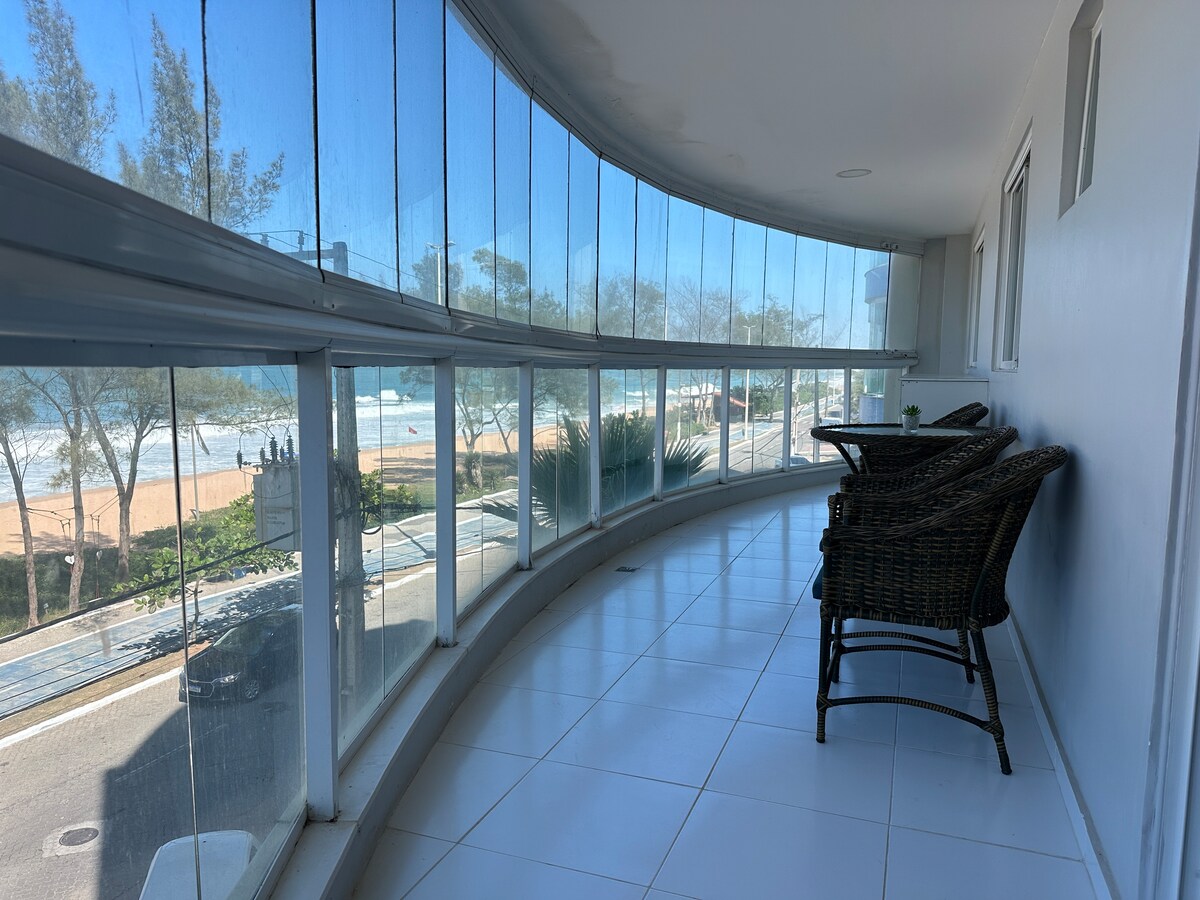 A large balcony features floor-to-ceiling glass curtains, offering unobstructed views of the beach. Two wicker chairs are positioned for comfortable seating. The tiled floor reflects natural light, creating a bright and open atmosphere. The surroundings include greenery and a glimpse of the ocean.