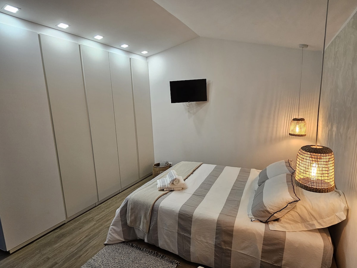 A cozy sleeping area features a spacious bed with soft striped bedding, complemented by a warm, neutral color palette. A wall-mounted television is visible, along with built-in wardrobes providing storage. Ambient lighting is created by woven pendant lamps on either side of the bed.