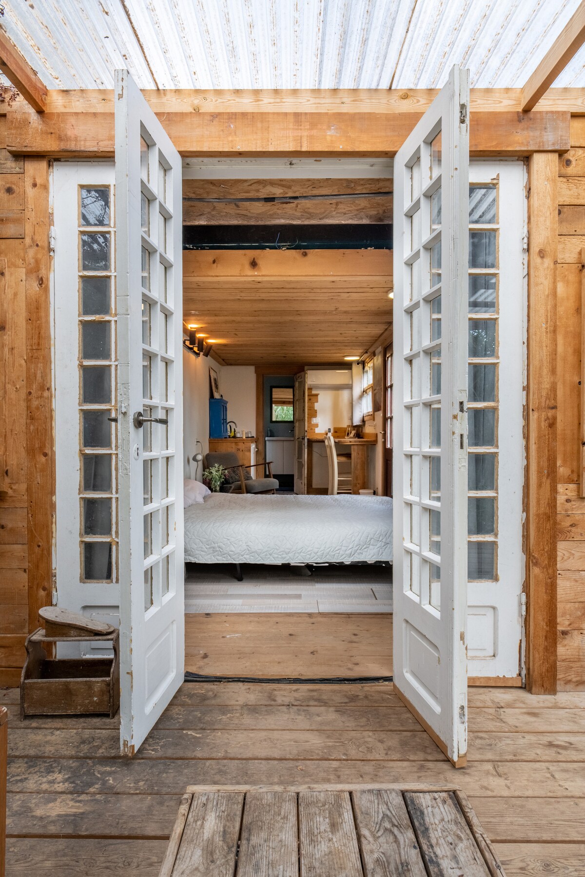 Double doors with divided glass panels open into a warm wooden interior, where a cozy sleeping area features a spacious bed. Natural light filters through the doors, highlighting the wooden flooring and simple decor of the cabin.
