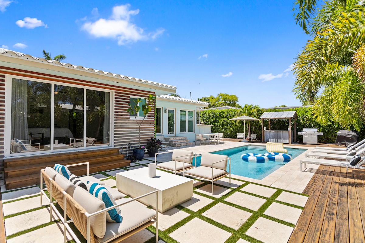 An outdoor area is featured, showcasing a heated pool surrounded by a spacious patio. Stylish lounge chairs and a shaded dining area are present, along with a modern outdoor grill. Lush greenery lines the perimeter, providing privacy and a serene atmosphere.