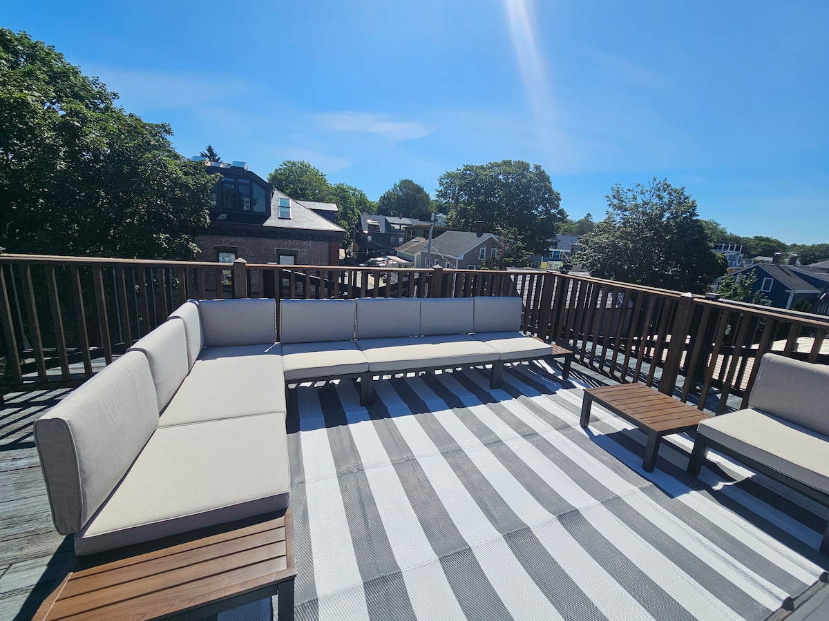 The rooftop deck is furnished with light-colored lounge seating arranged around a wooden table. A striped outdoor rug stretches across the space, while views of surrounding greenery and nearby buildings are visible in the background under a clear blue sky.