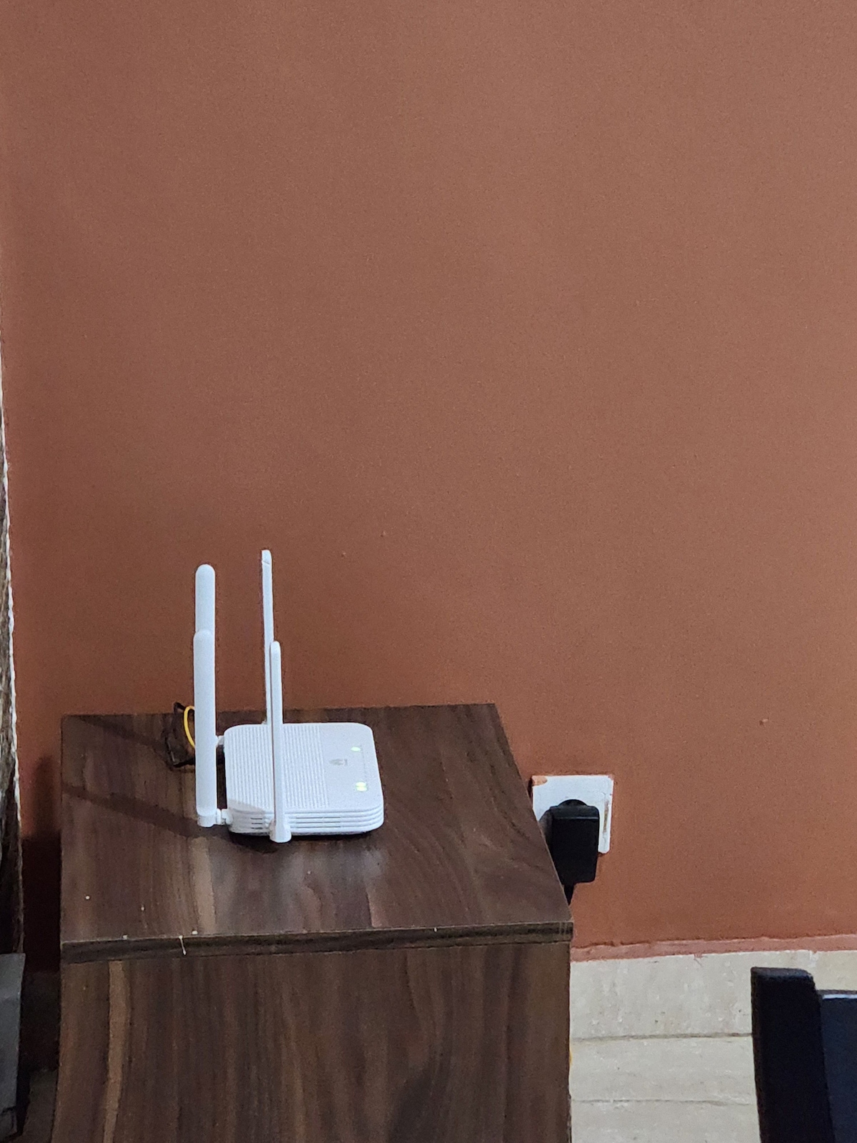 A wireless router is positioned on a wooden surface against a painted wall. The router features two antennae, ensuring connectivity throughout the apartment. An electrical outlet is visible nearby, adding to the utility of the space.