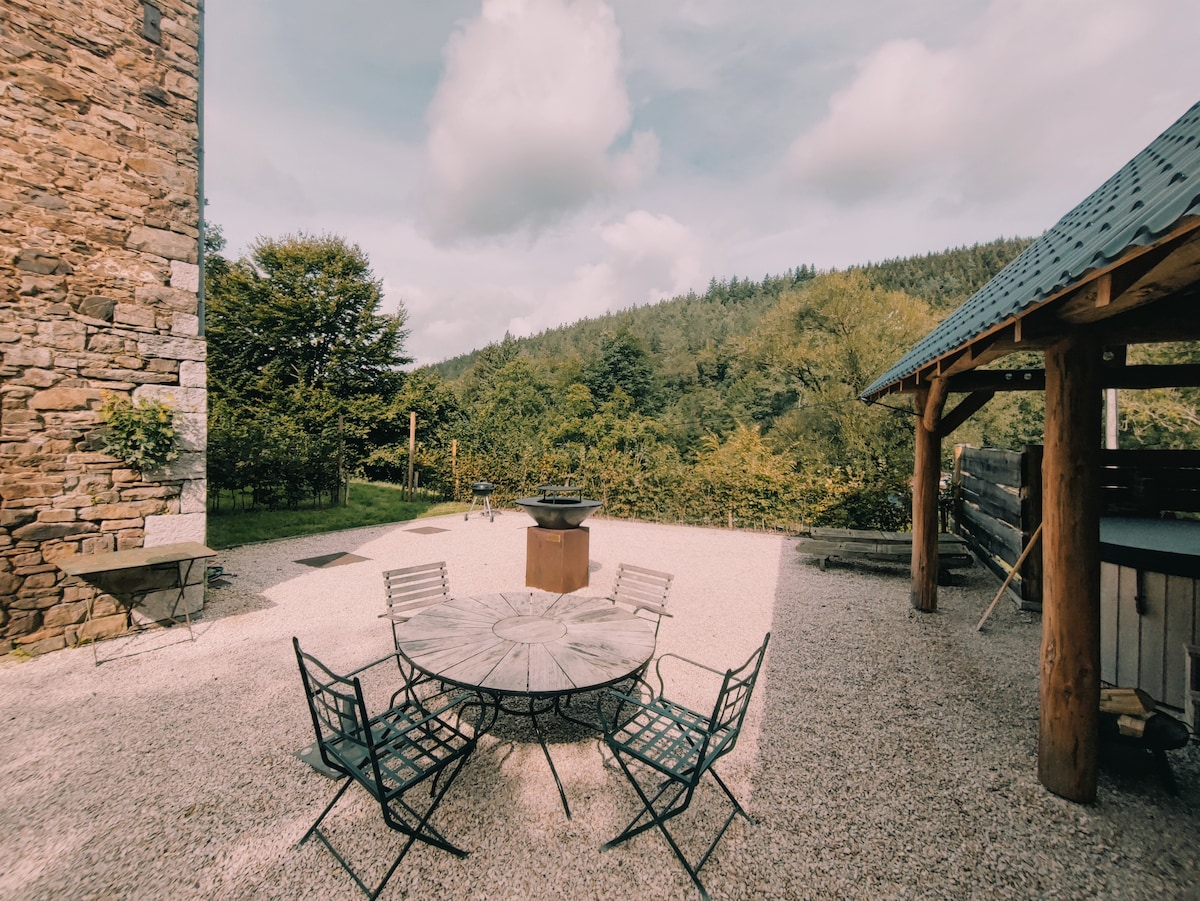 An outdoor terrace features a round wooden table surrounded by six metal chairs, set on gravel. In the background, lush greenery and rolling hills are visible, under a partly cloudy sky. A barbecue grill is positioned nearby, enhancing the inviting outdoor space.