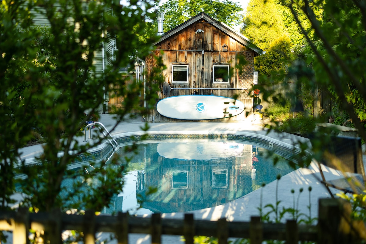 An inviting inground pool is framed by lush green foliage, reflecting the sunlight. A rustic wooden cabin with windows stands in the background, featuring a surfboard resting against its weathered exterior. The surrounding area includes a well-tended garden that enhances the serene ambiance.
