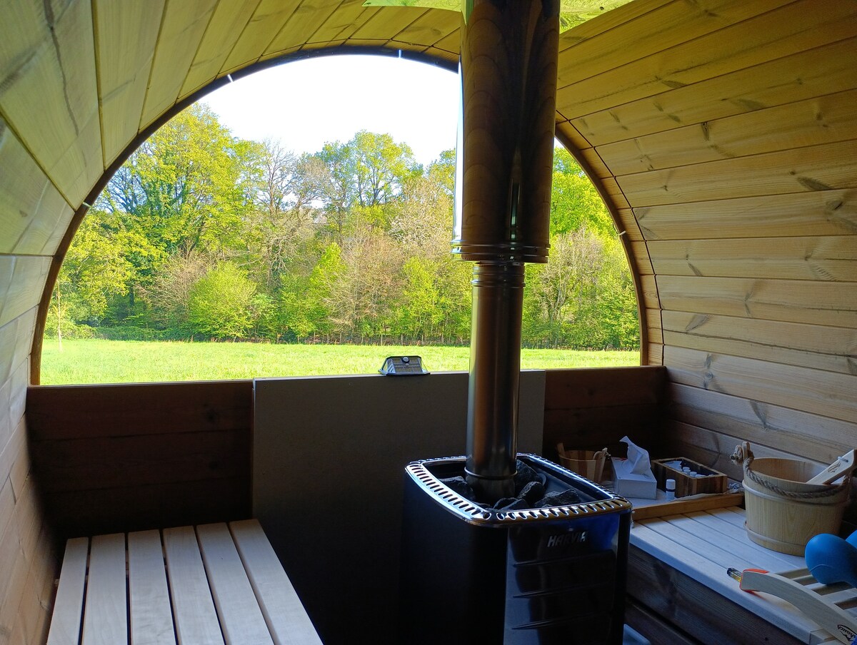 The image captures the interior of a wooden sauna, featuring a circular window that frames a lush green forest view. A sleek metal stove is positioned centrally, surrounded by wooden benches, creating a cozy and functional space for relaxation.