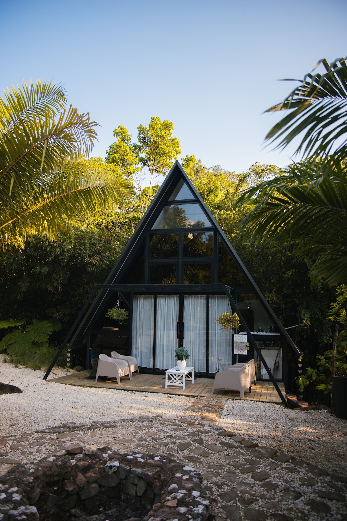 An A-frame cabin is surrounded by lush greenery, showcasing large front windows that allow natural light to fill the interior. A small patio area features two chairs and a table, providing a cozy space to enjoy the outdoor setting.