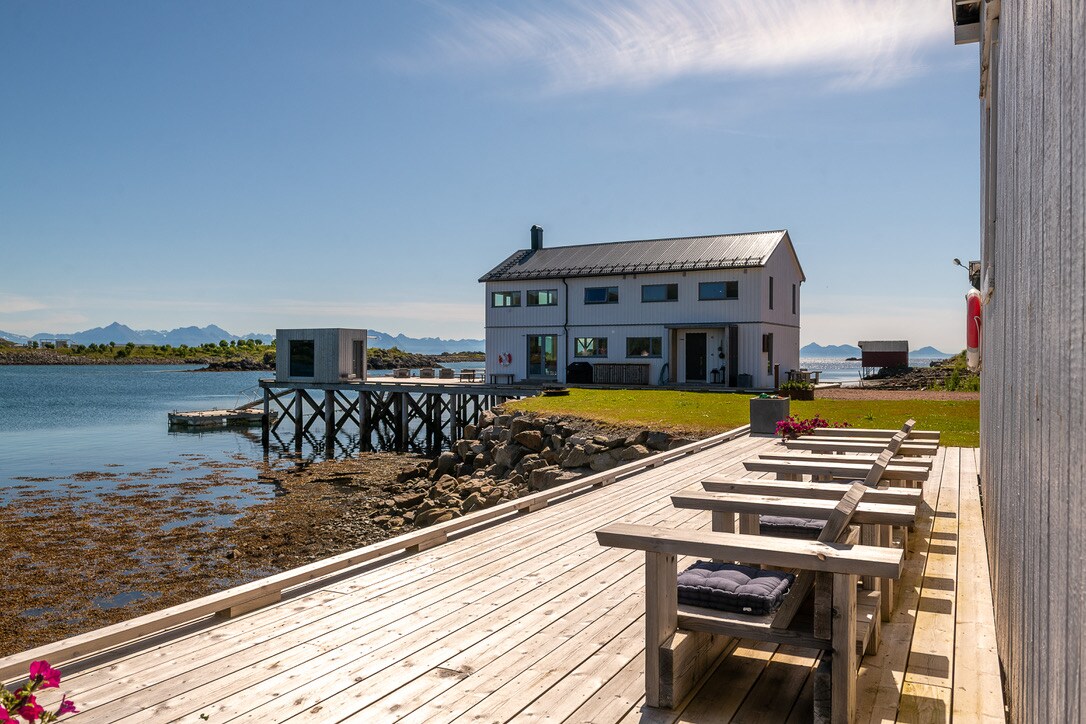 A scenic view of a modern coastal house positioned beside the water. Wooden benches are arranged along a spacious deck, providing seating for relaxation. The surrounding landscape features calm water and distant mountains, creating a serene atmosphere.