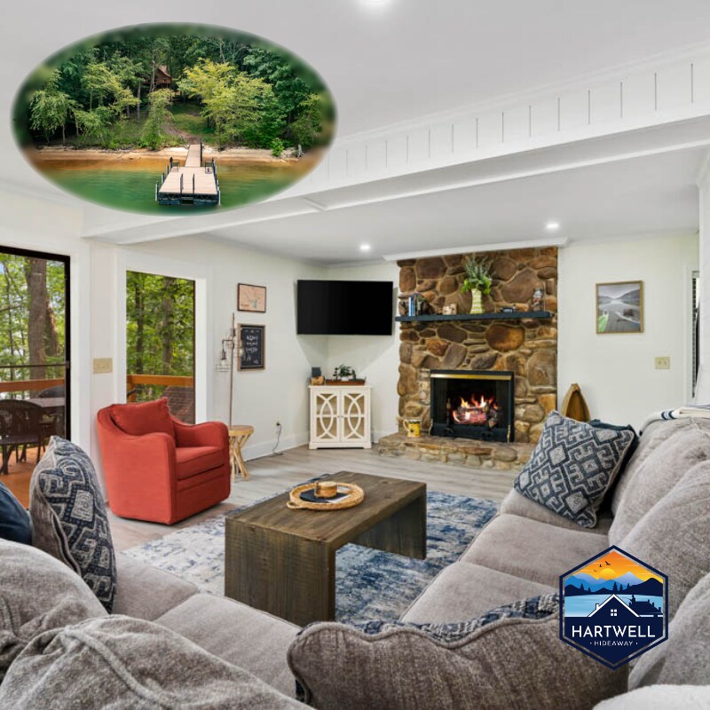 The living room showcases a comfortable sectional sofa and an accent chair, with a stone fireplace as a focal point. A large flat-screen TV is mounted above the mantle. Views of the lake are visible through the large windows, which connect to the outdoor space.